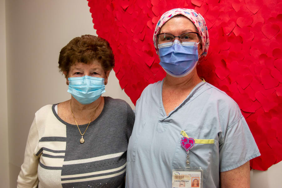A woman stands beside a nurse in a hospital against a backdrop of cutout hearts. They are wearing medical masks and looking at the camera.