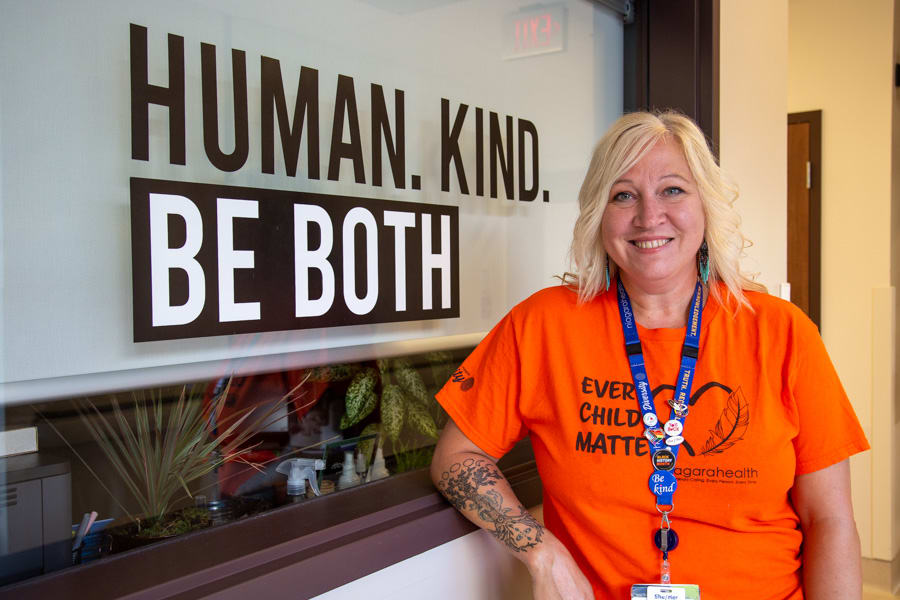 Debby Schaubel in an orange t-shirt stands in front of a sign that says Human. Kind. Be Both.